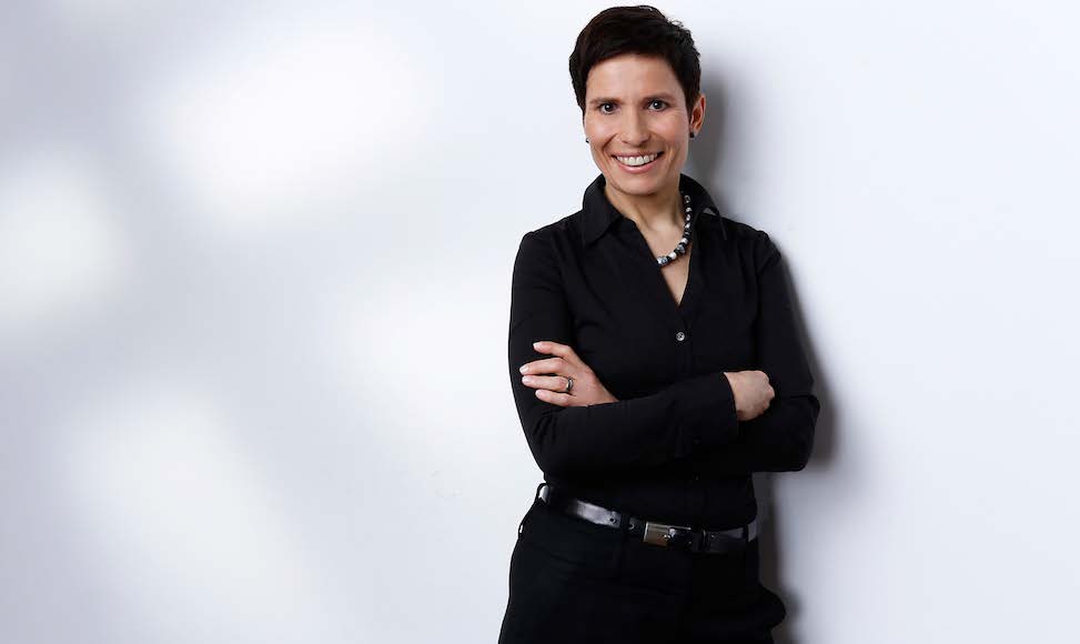 Stefanie Groos steht vor einer weißen Wand mit Schattenspiel im Hintergrund. Sie trägt kurze dunkle Haare und dunkle Kleidung. Ihre Arme sind vor ihrem Oberkörper verschränkt. Sie lächelt in die Kamera.